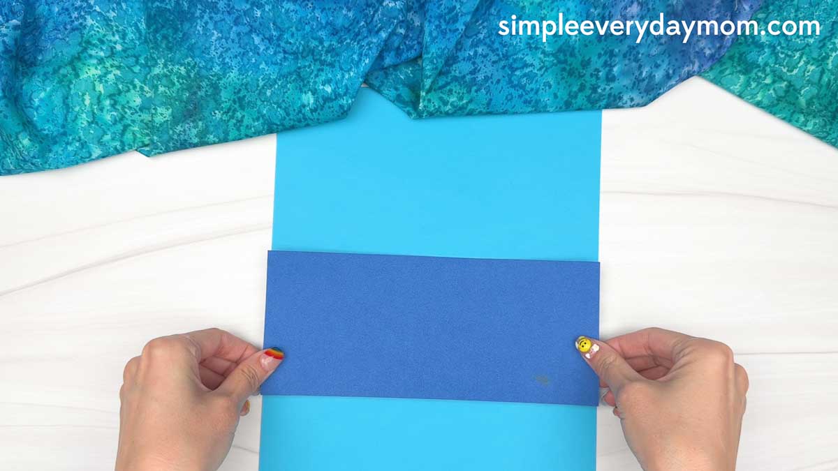 Hands holding a dark blue paper strip over a light blue sheet to assemble a reindeer summer craft.