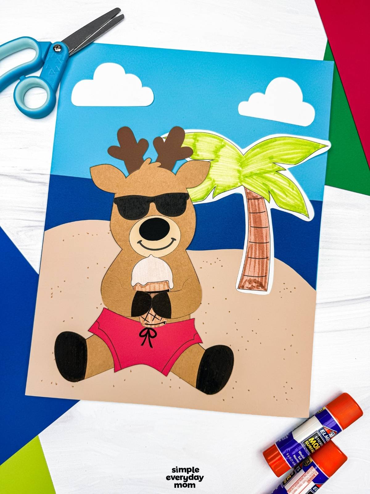 A completed craft featuring a reindeer in sunglasses and green swim trunks holding an ice cream cone on a beach scene background with a palm tree and clouds.
