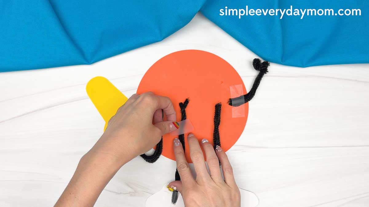 Hands taping pipe cleaner legs onto the back of a basketball Father’s Day paper craft.