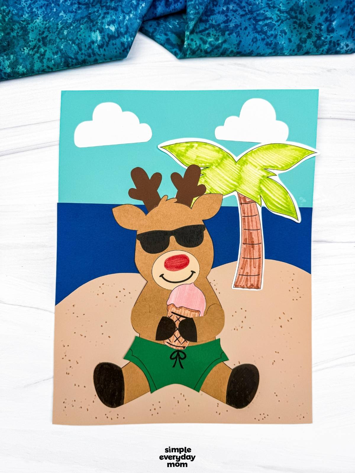 A completed craft featuring a reindeer in sunglasses and green swim trunks holding an ice cream cone on a beach scene background with a palm tree and clouds.