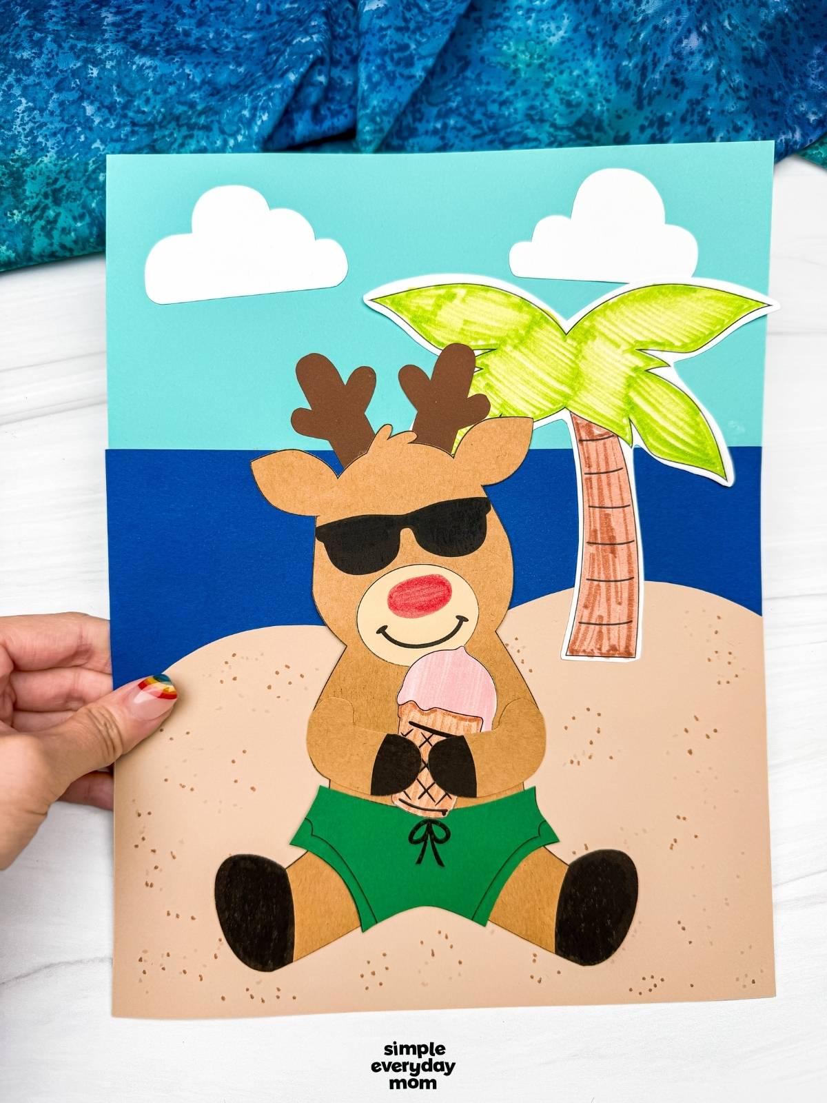 Hands holding a completed reindeer summer craft on a beach scene background with a palm tree, clouds, sunglasses, and an ice cream cone.