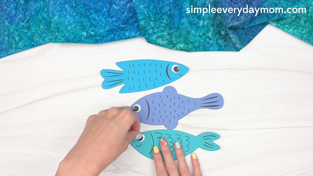 Hands placing googly eyes on colorful paper fish pieces for a Father’s Day fishing craft.
