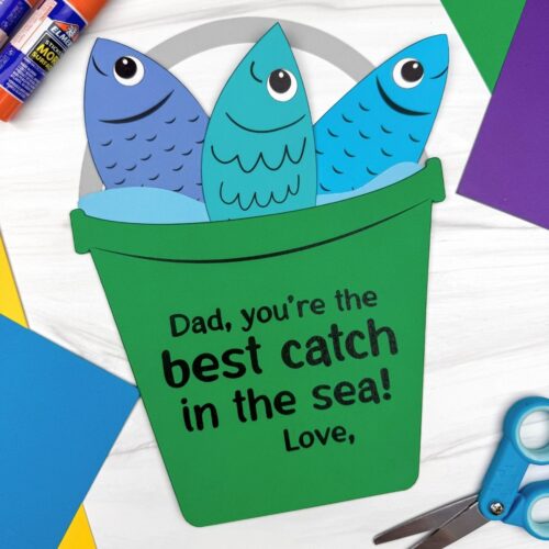 A completed Father’s Day fishing bucket craft with colorful paper fish surrounded by craft supplies and cardstock paper.