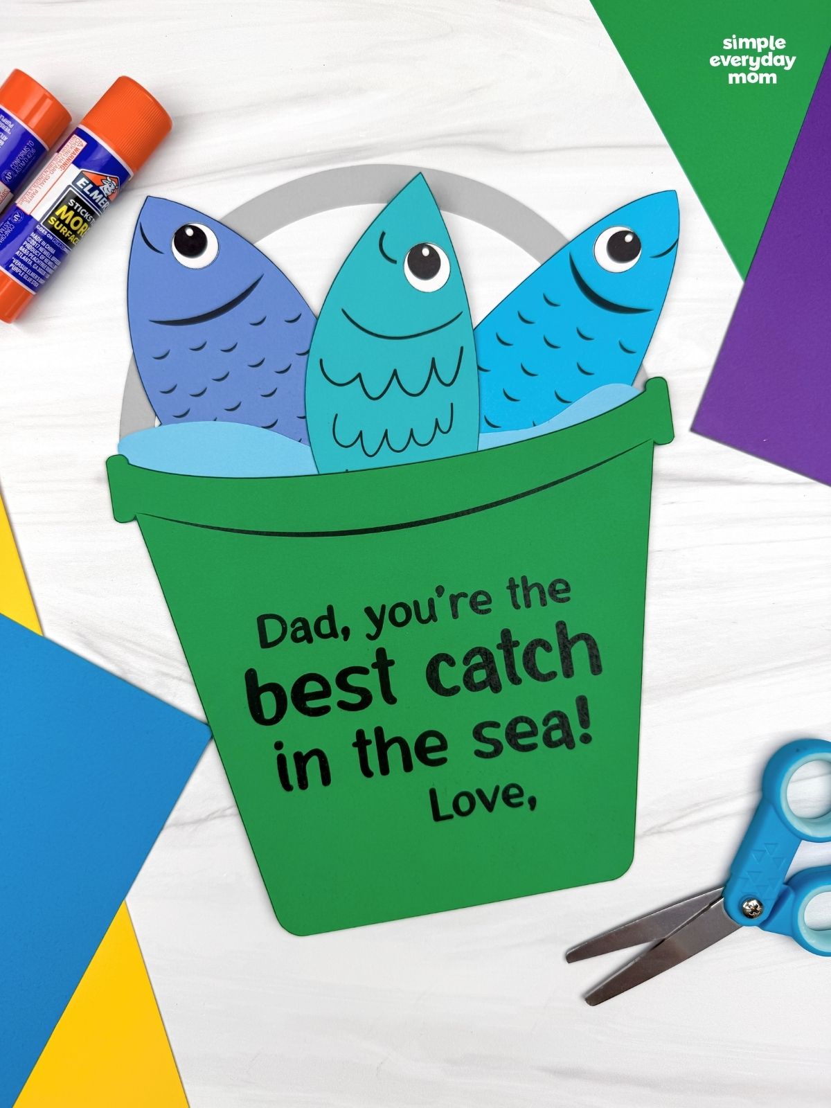 A completed Father’s Day fishing bucket craft with colorful paper fish surrounded by craft supplies and cardstock paper.