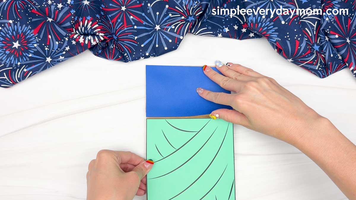 Hands attaching the Statue of Liberty body cutout to a brown paper bag with a blue background piece already in place.
