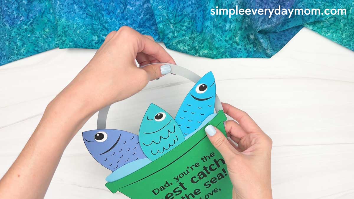 Hands attaching a handle to a completed Father’s Day fishing bucket craft with colorful paper fish.