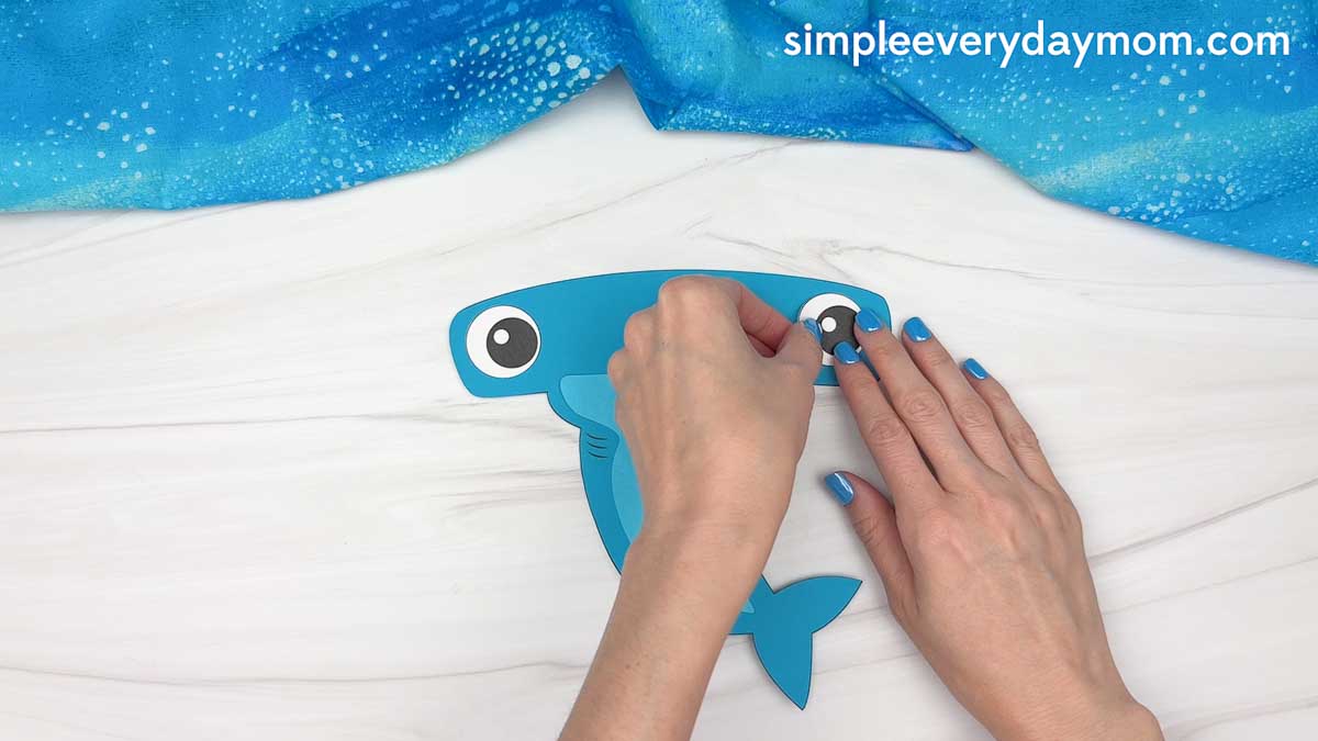 Hands attaching a paper eye to a blue hammerhead shark craft during assembly.