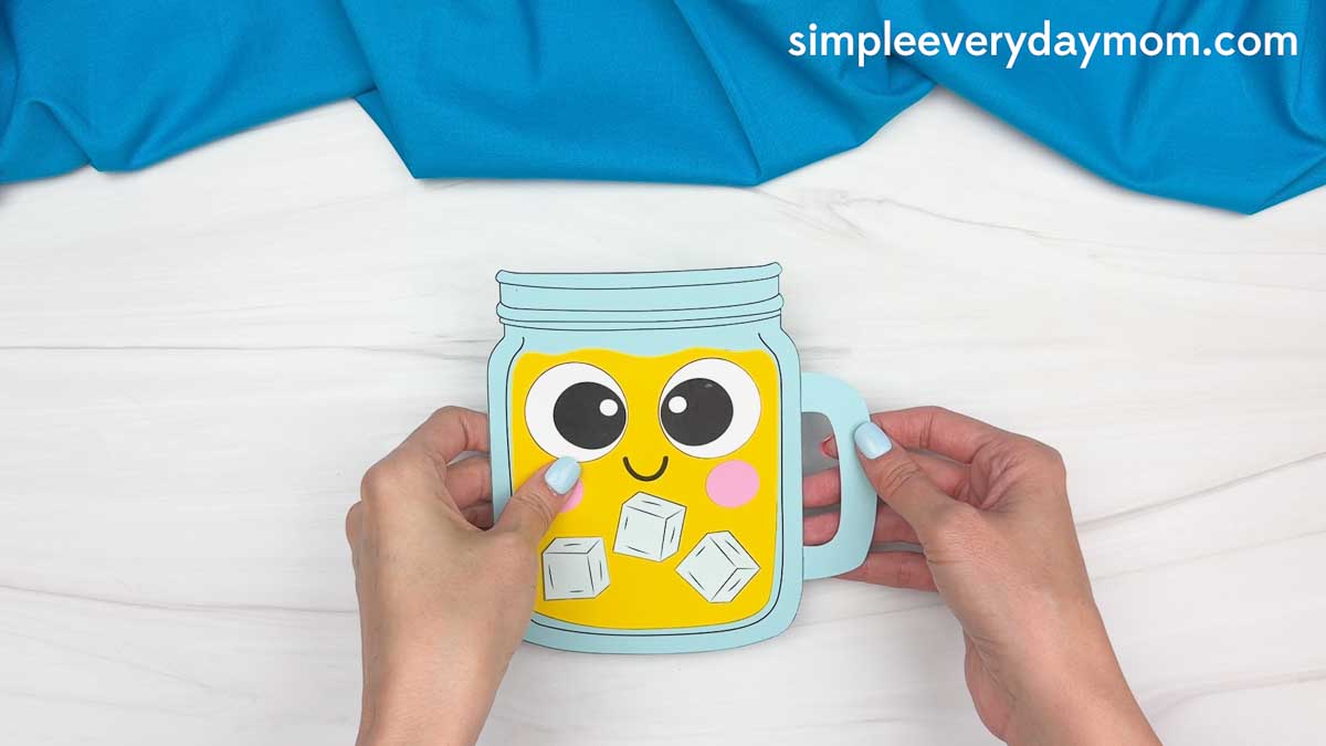 Hands holding a completed lemonade jar craft with a smiling face, pink cheeks, and paper ice cubes