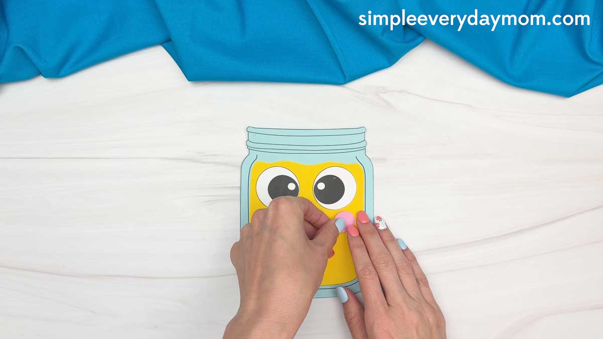 Hands attaching pink cheeks to a smiling lemonade jar craft with large eyes.