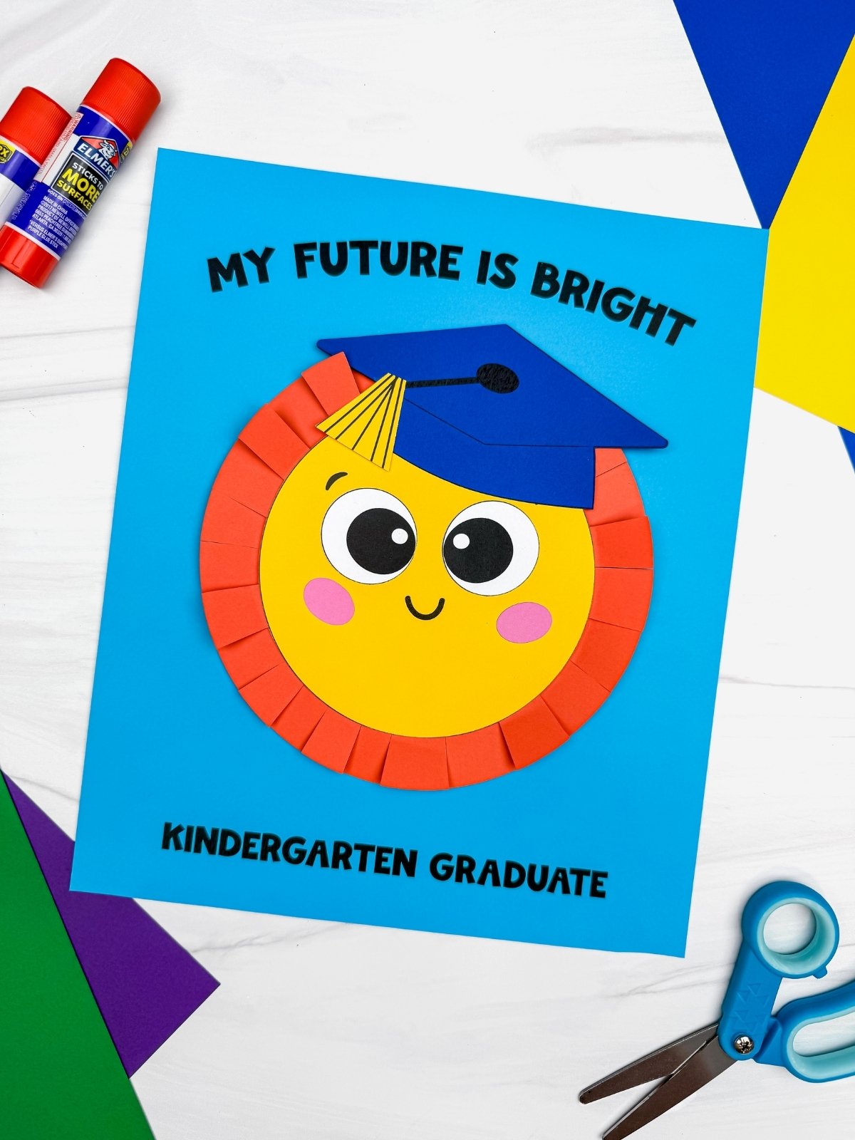 A completed craft sun graduation card with a smiling sun wearing a blue graduation cap surrounded by colorful craft supplies on a white table