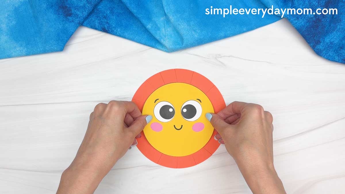 Hands gluing a smiling yellow sun face onto orange paper sun rays for a graduation craft activity