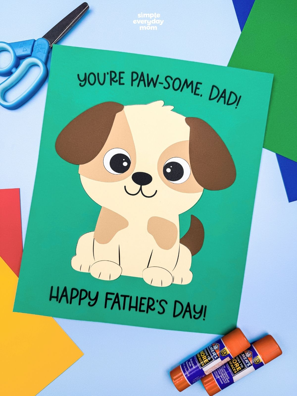 A completed craft featuring a puppy Father’s Day card with colorful paper sheets, scissors, and glue sticks for a kids craft activity