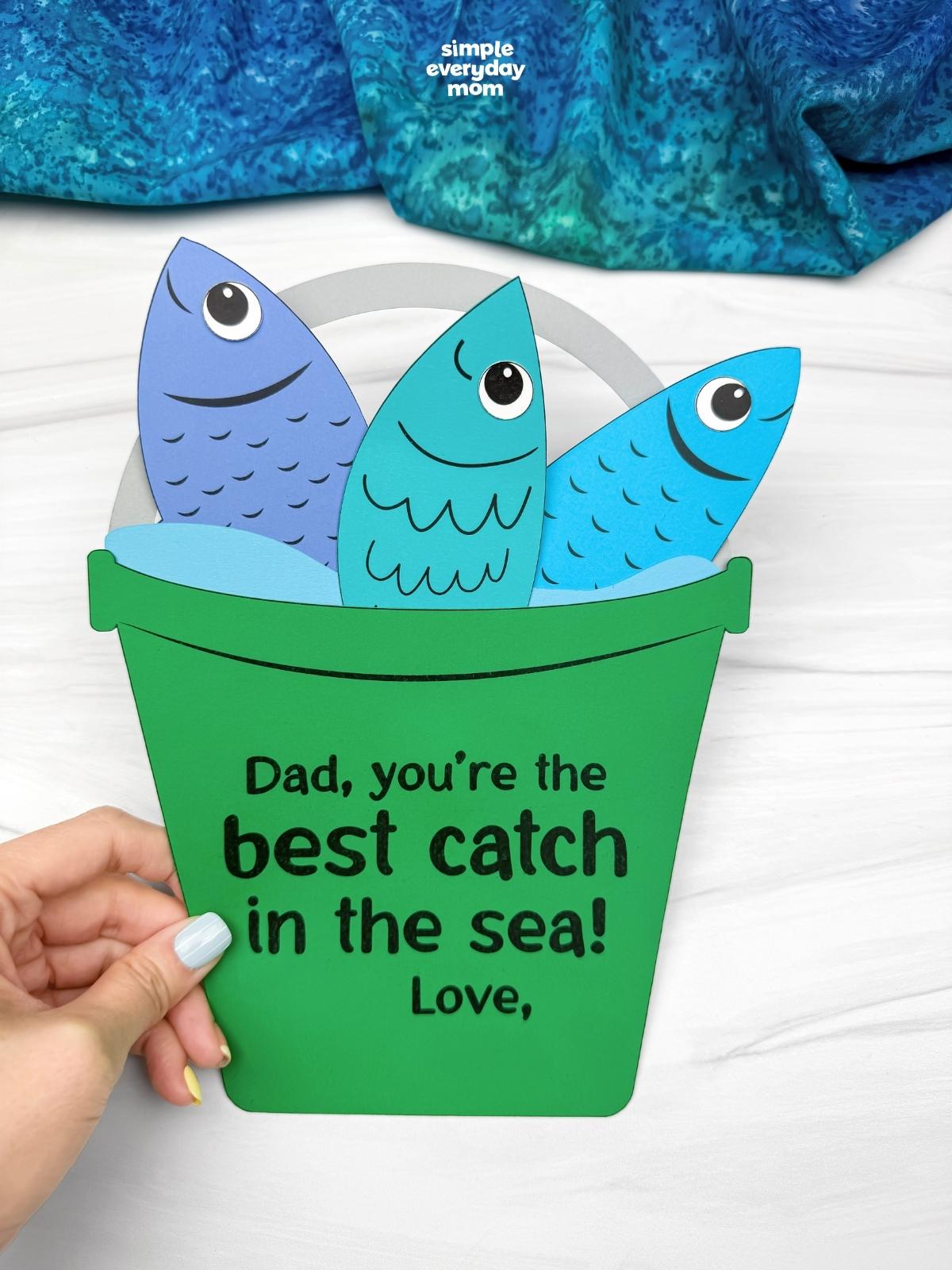 Hands holding a completed Father’s Day fishing bucket craft with colorful paper fish and a best catch in the sea message.
