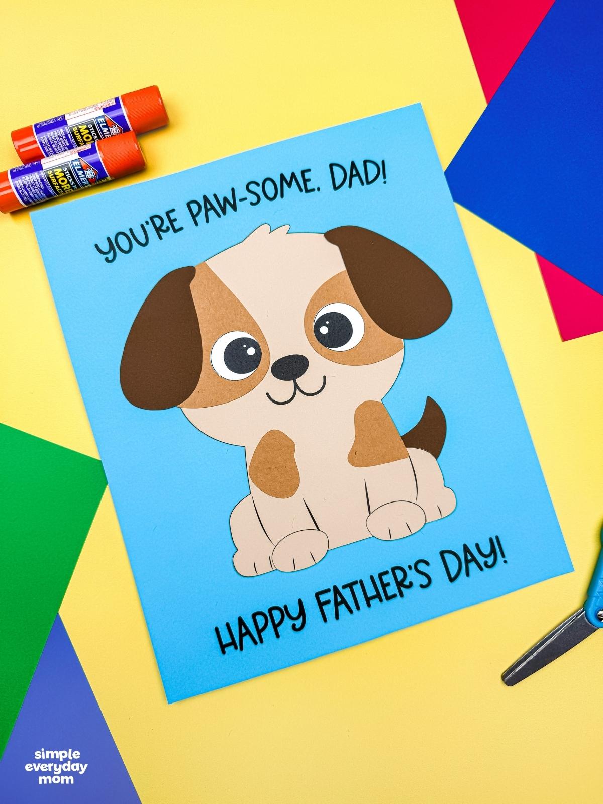 A completed craft featuring a puppy Father’s Day card with colorful paper supplies and glue sticks on a craft table