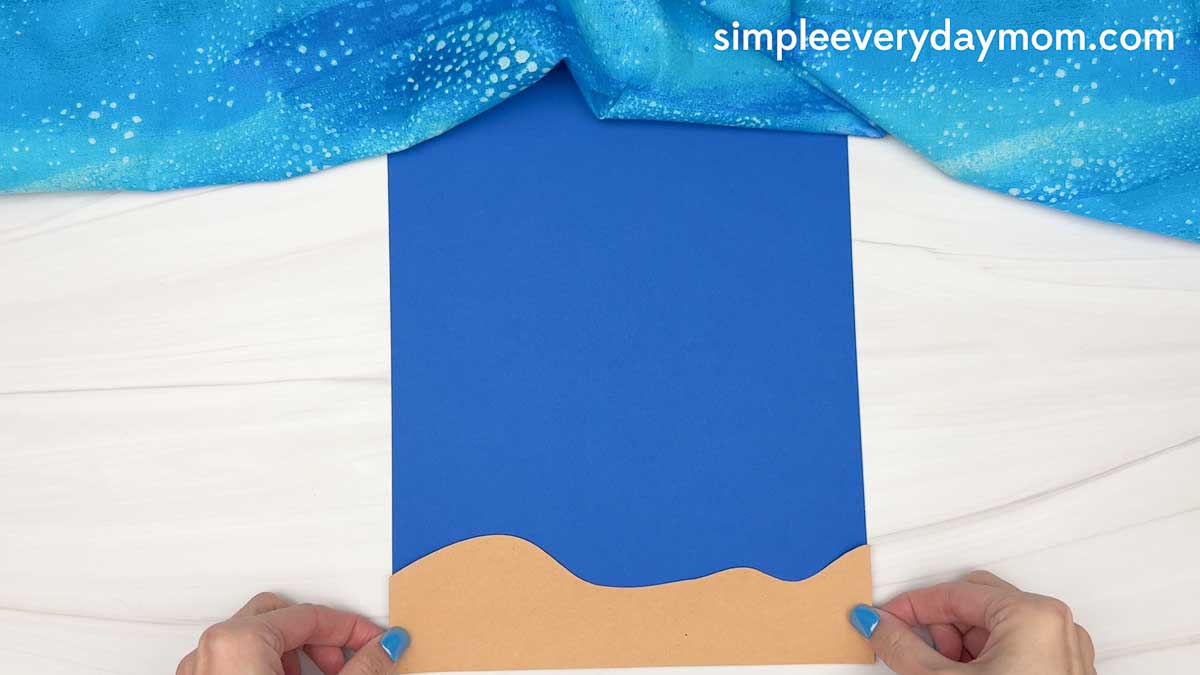 Hands positioning a tan paper sand piece onto a blue ocean background for a hammerhead shark craft.