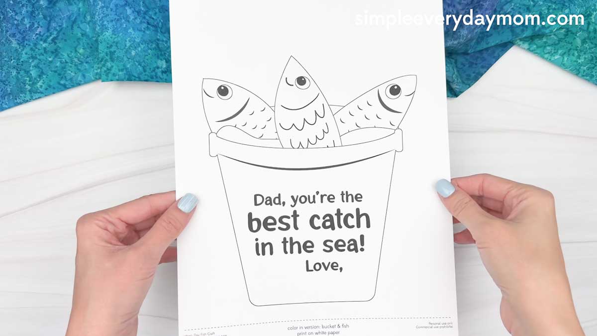 Hands holding a printable Father’s Day fishing craft template with fish in a bucket and “best catch in the sea” message.
