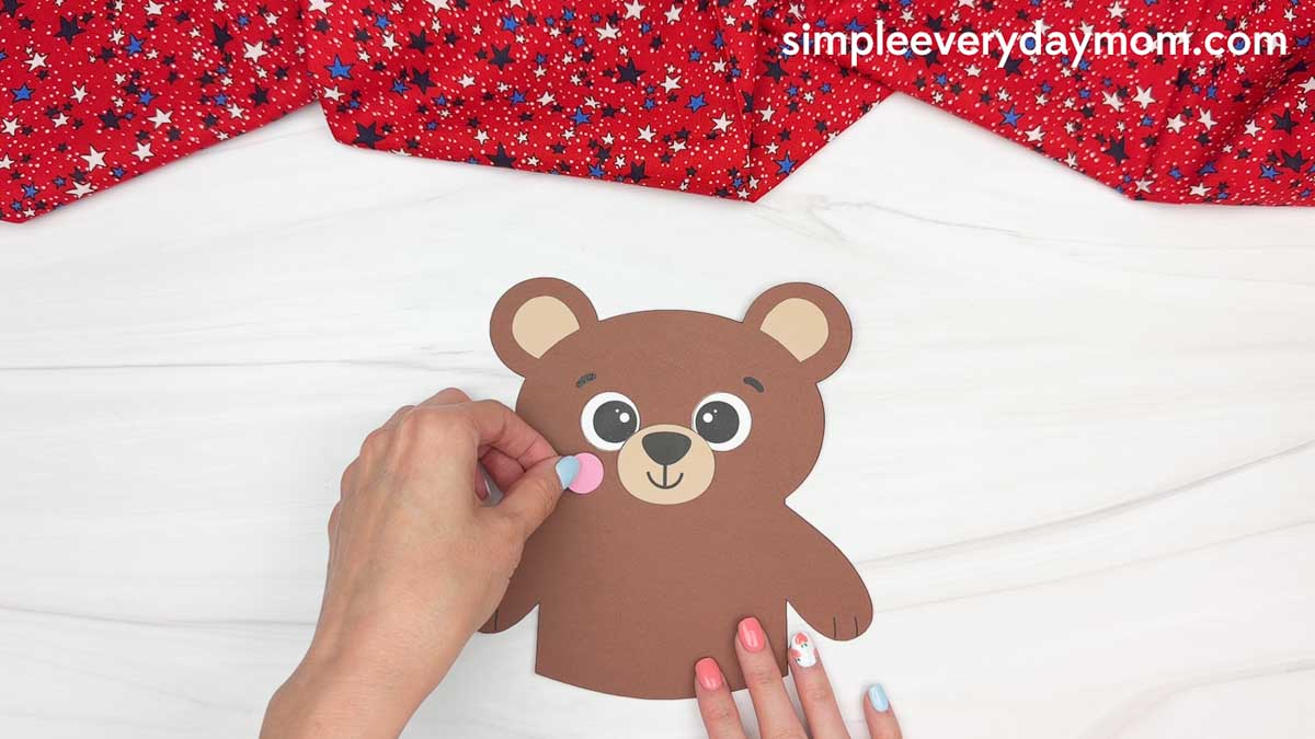 Hands attaching a pink cheek piece to a brown teddy bear paper craft for a patriotic teddy bear activity