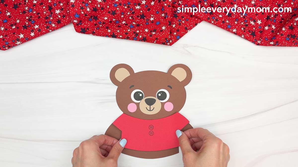Hands holding a red shirt piece on a brown teddy bear paper craft for a patriotic teddy bear activity