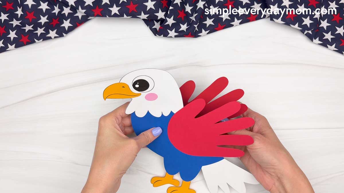 Hands holding a completed American eagle handprint craft while attaching a red handprint wing to the side.