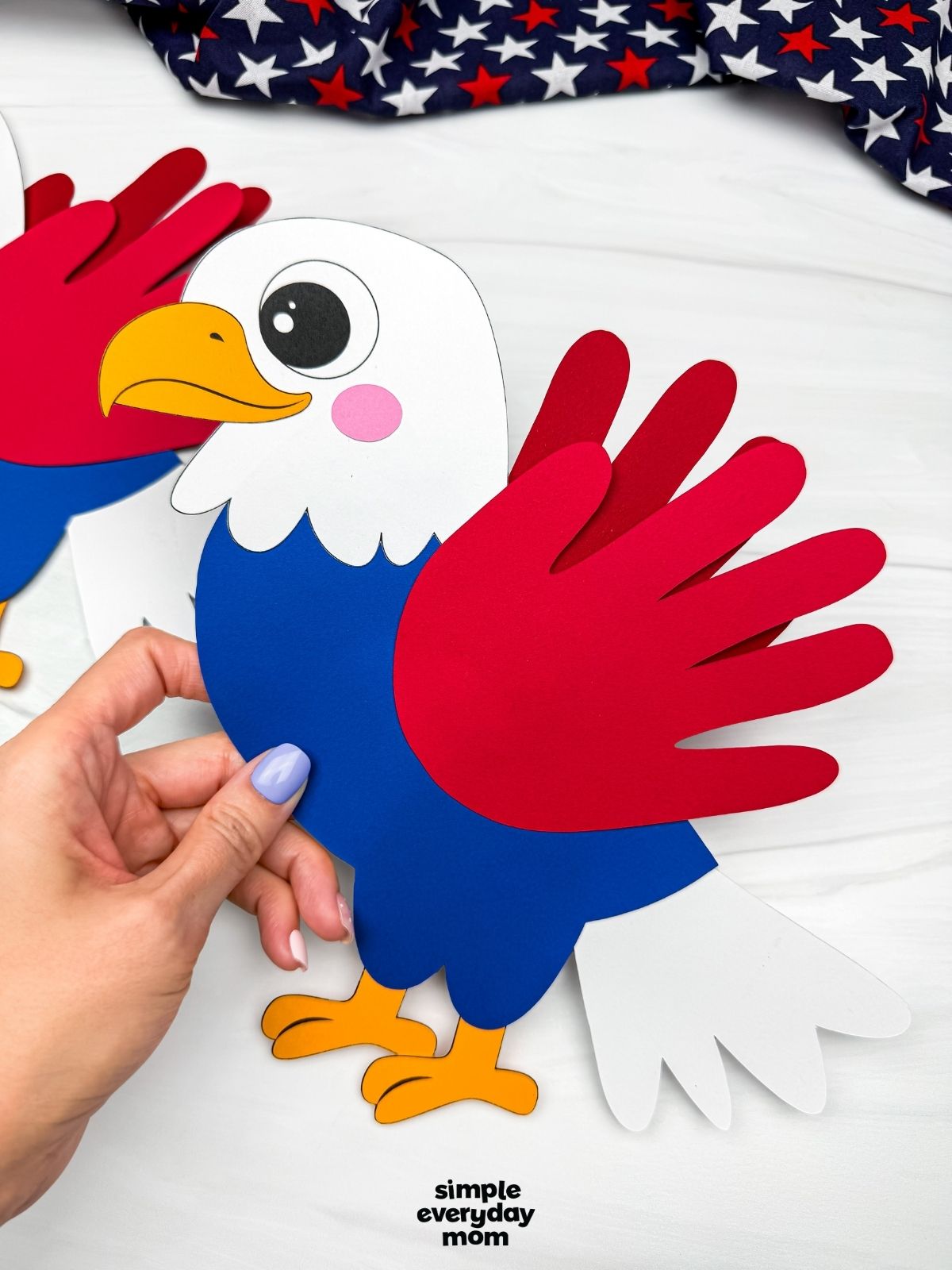 Hands holding a completed American eagle handprint craft with red handprint wings, white tail feathers, and a blue body.