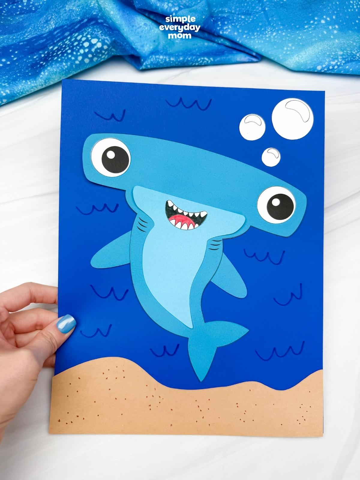 Hands holding a completed hammerhead shark craft on a blue underwater background with bubbles and a sandy ocean floor.