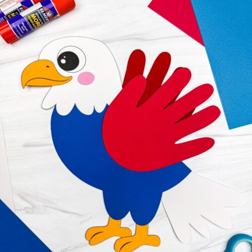 A completed craft showing an American eagle handprint craft surrounded by colorful paper, glue sticks, and scissors.