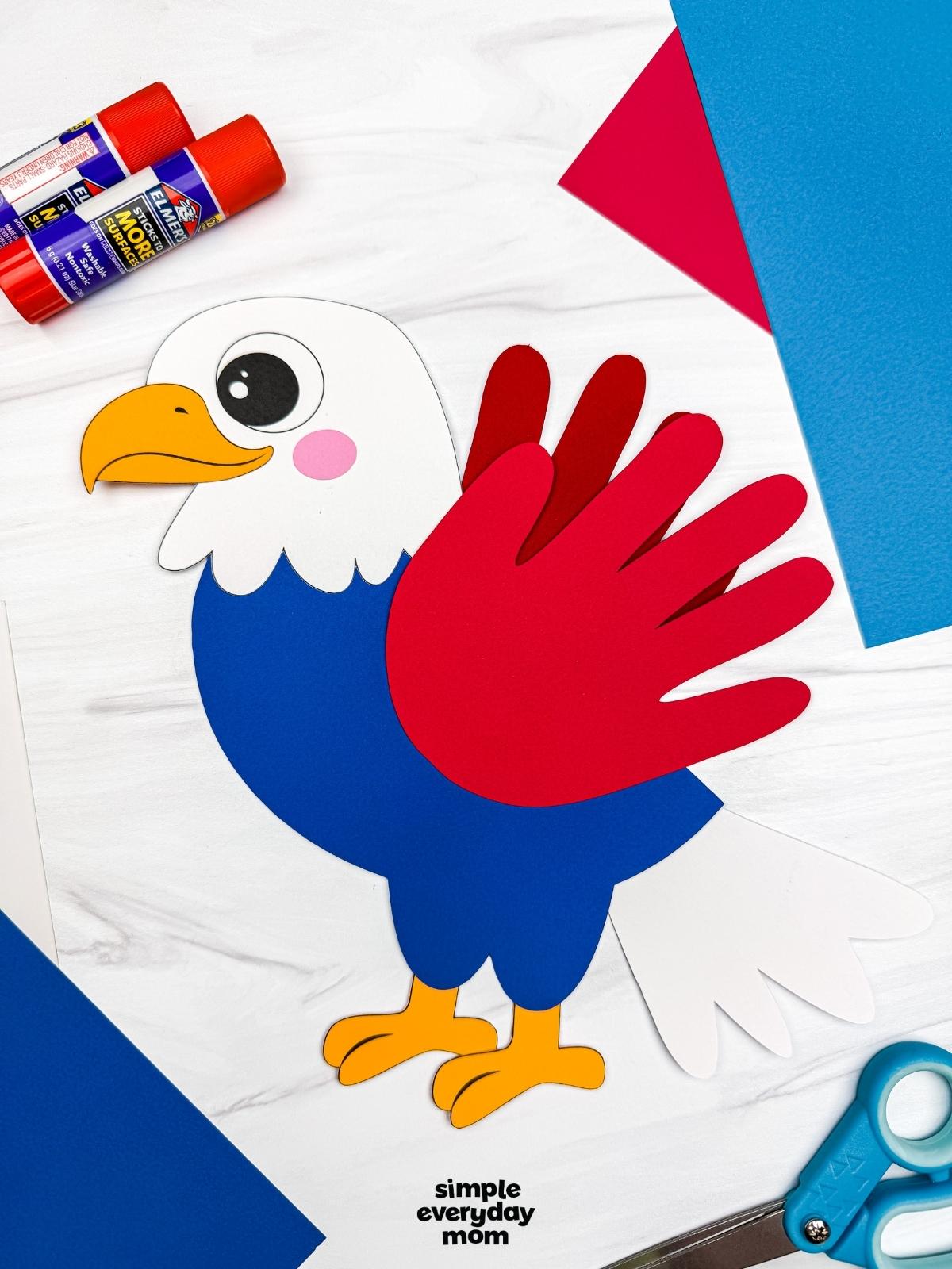A completed craft showing an American eagle handprint craft surrounded by colorful paper, glue sticks, and scissors.