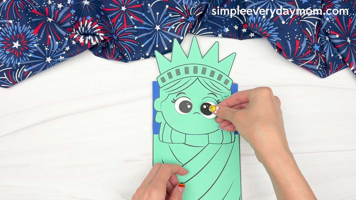 Hands attaching a large eye piece to the Statue of Liberty paper bag puppet craft face.