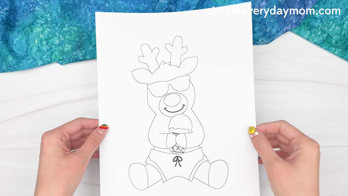 Hands holding a printed reindeer summer craft template featuring a reindeer in sunglasses and swim trunks holding an ice cream cone.