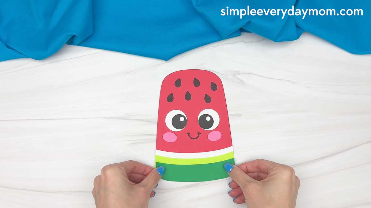 Hands holding a completed summer popsicle craft with a smiling watermelon face and layered green and white stripes.