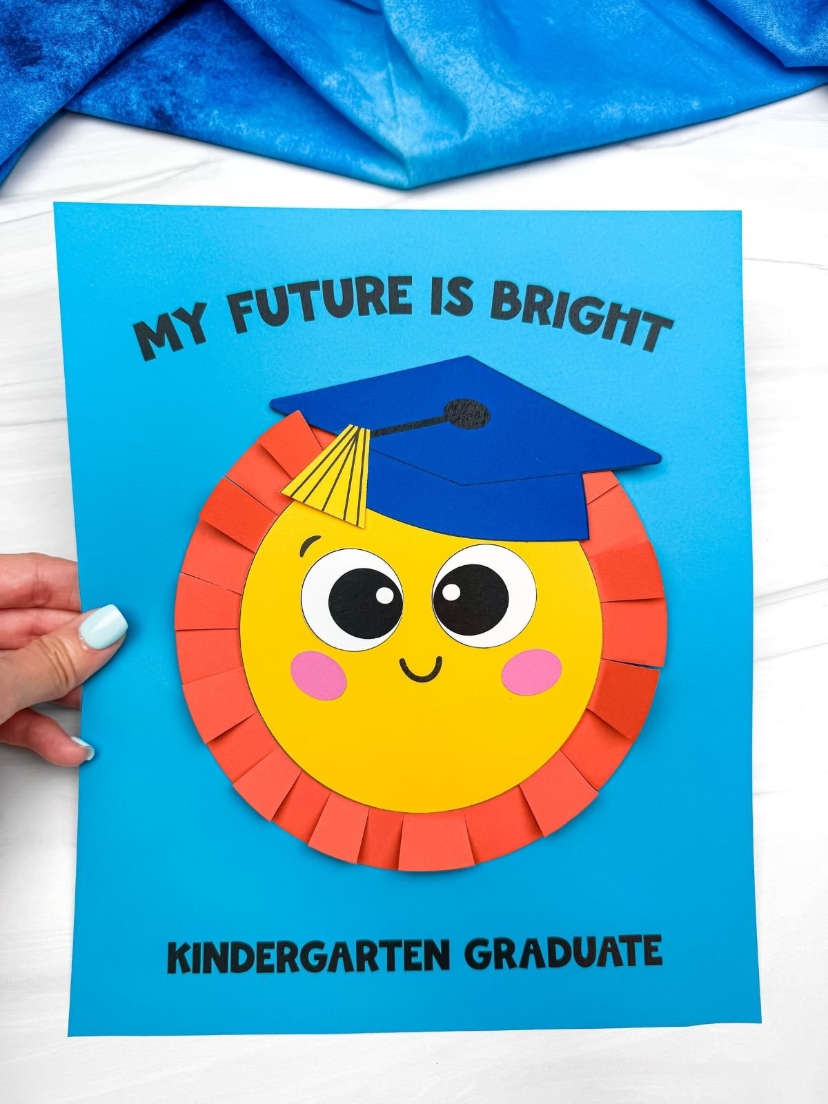 Hands holding a completed sun graduation craft card with a smiling sun wearing a blue graduation cap and “My Future Is Bright” text on blue paper