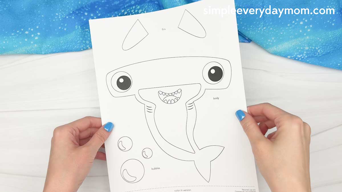 Hands holding a printed hammerhead shark craft template with cutout pieces for fins, body, and bubbles.