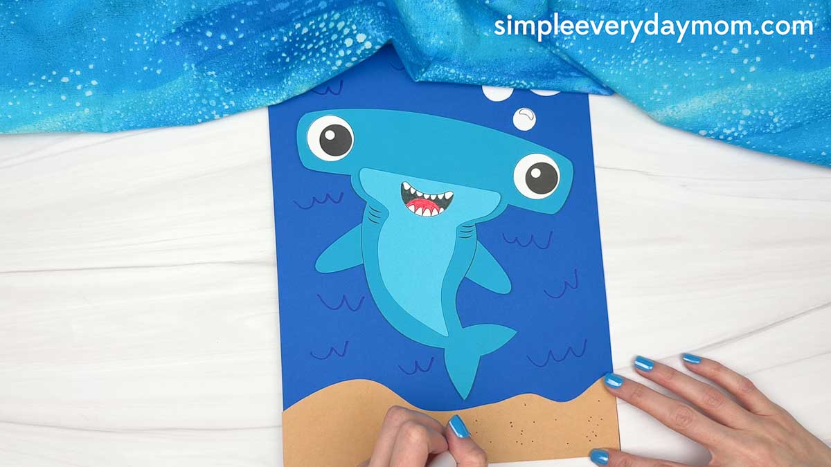 Hands adding details to a completed hammerhead shark craft on an underwater ocean scene with bubbles and a sandy seafloor.