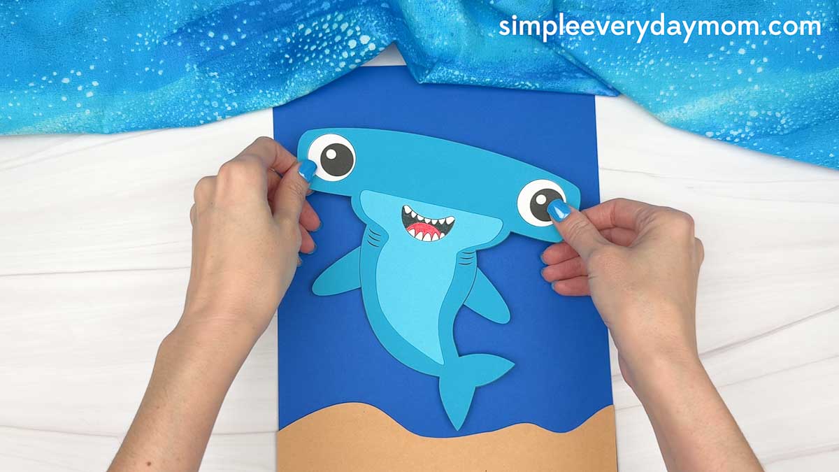 Hands placing a completed hammerhead shark craft onto a blue ocean background with a sandy seafloor.