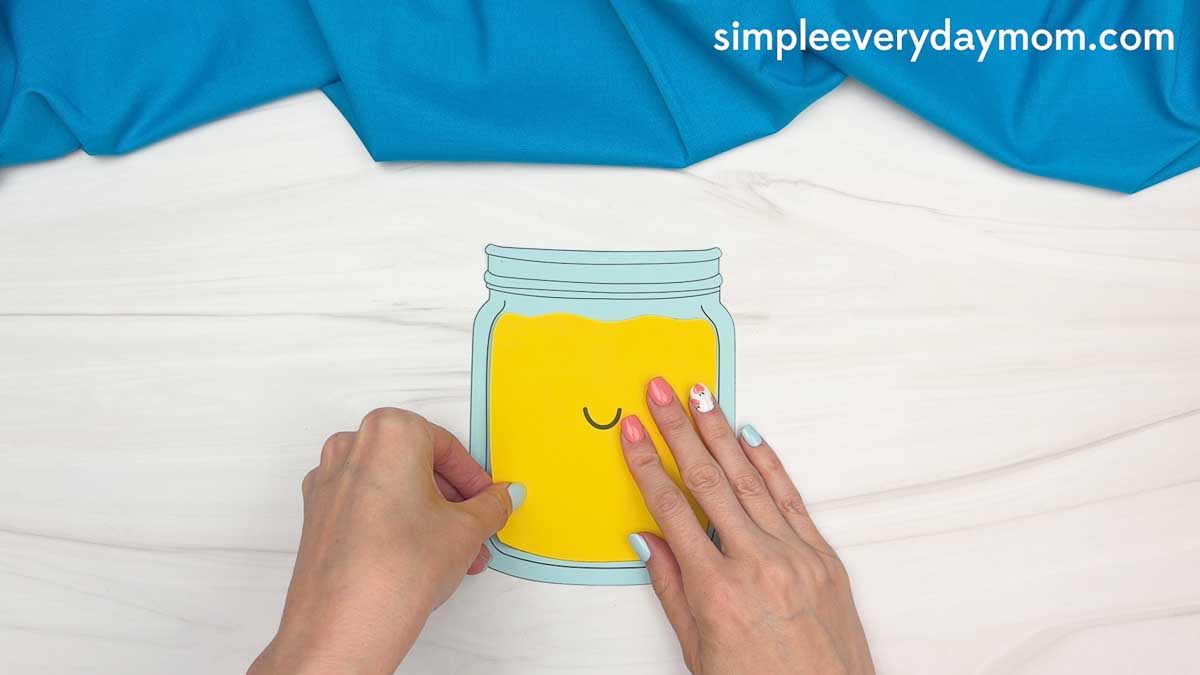 Hands placing a smiling lemonade jar craft cutout on a white table.