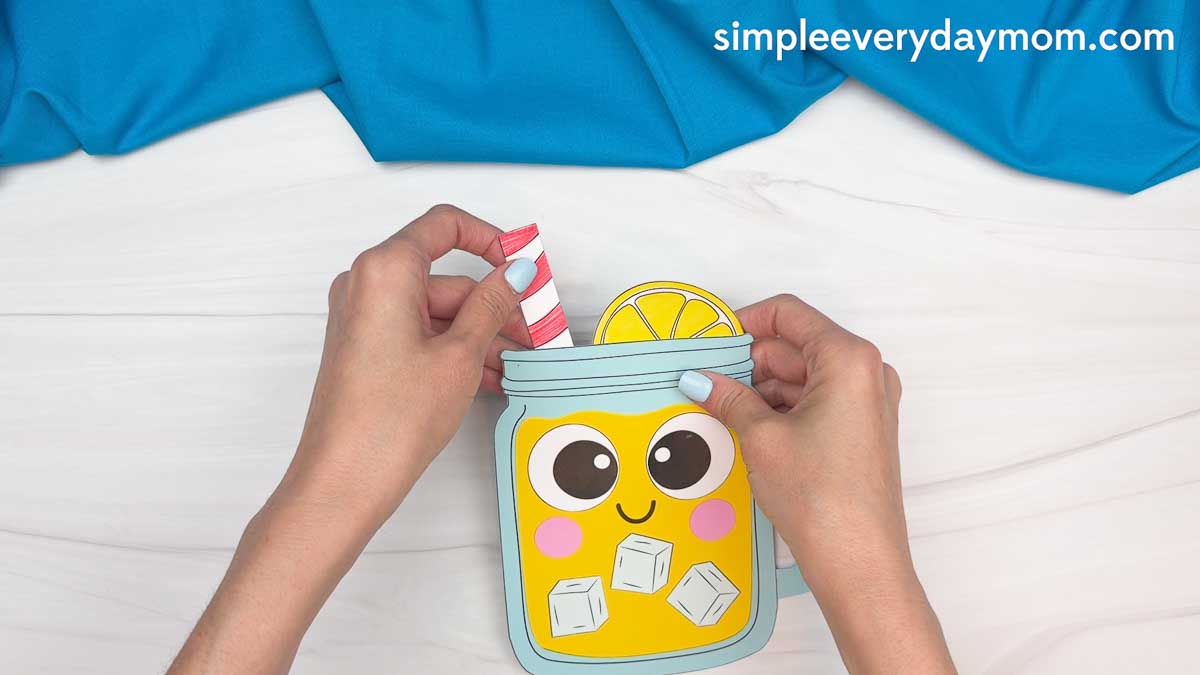 Hands placing a striped paper straw into a completed lemonade jar craft with a lemon slice and ice cubes.
