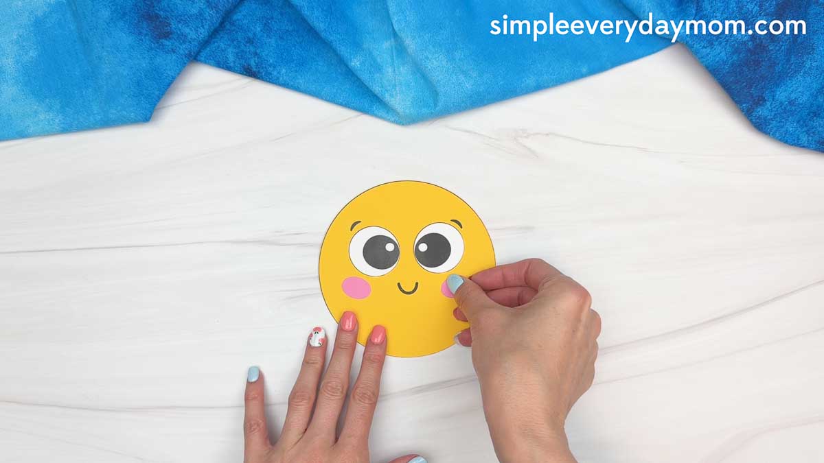 Hands attaching pink cheeks to a smiling yellow sun cutout for a graduation craft project