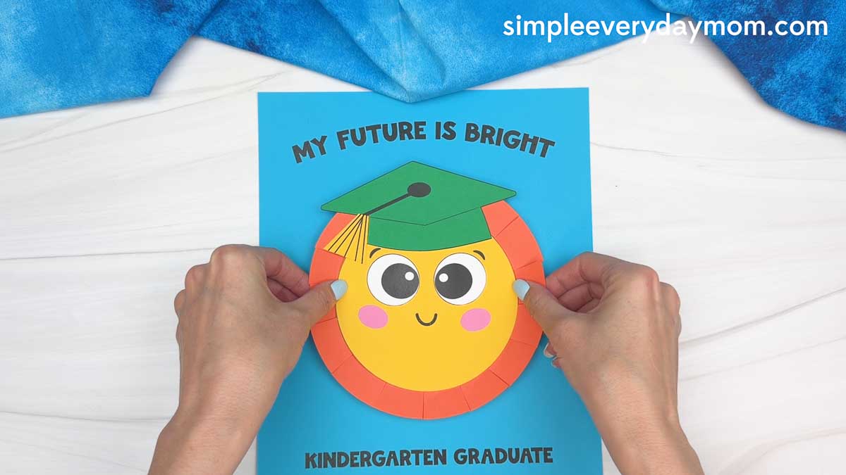Hands placing a completed sun graduation craft onto a blue background with “My Future Is Bright” and “Kindergarten Graduate” text