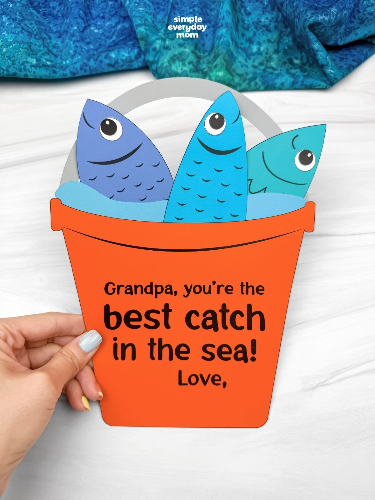 Hands holding a completed Grandpa fishing bucket craft with colorful paper fish and a best catch in the sea message.
