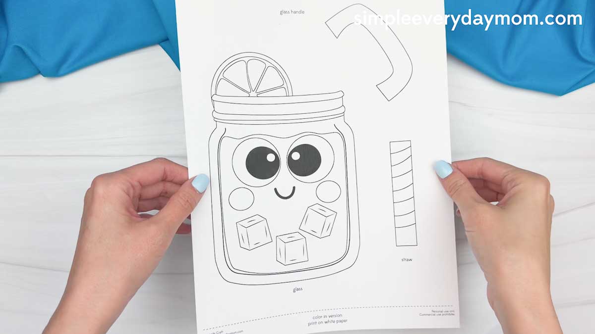 Hands holding a printable lemonade craft template with a smiling mason jar, straw, lemon slice, and ice cubes.
