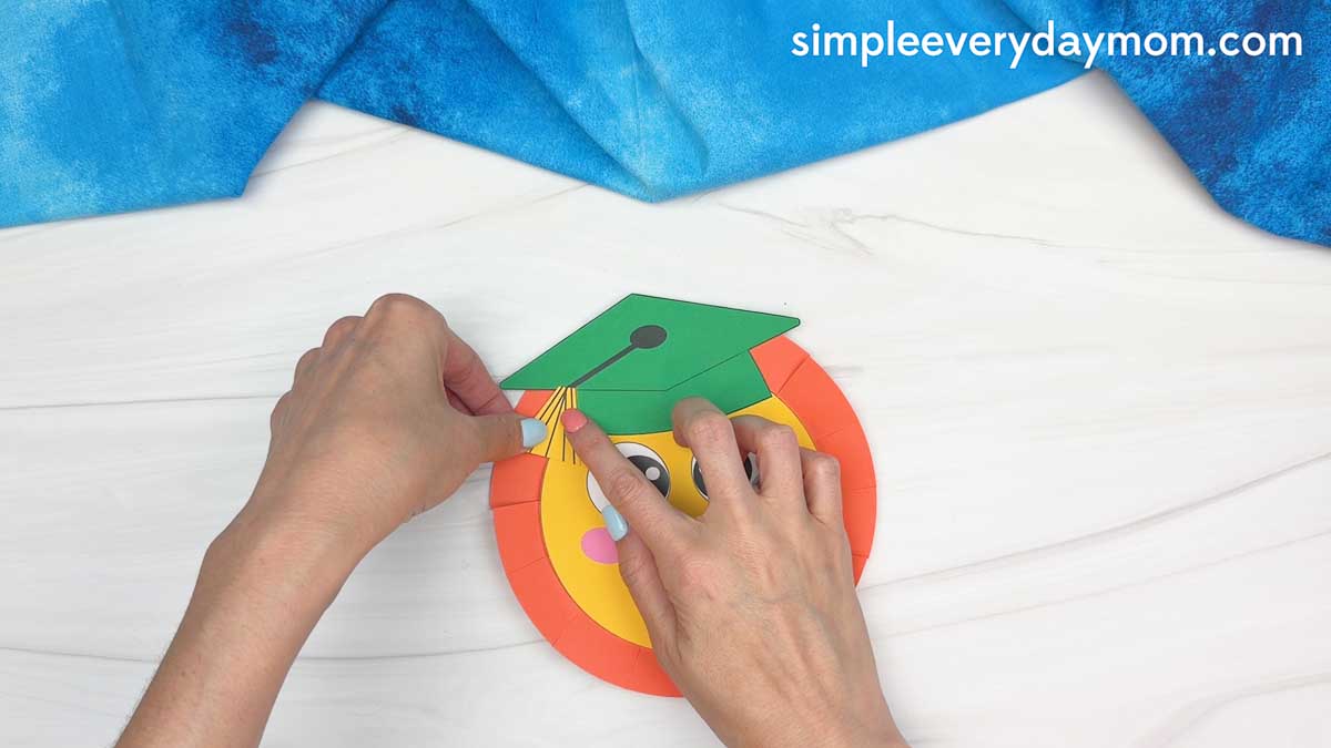 Hands gluing a yellow paper tassel onto a green graduation cap on a smiling sun craft
