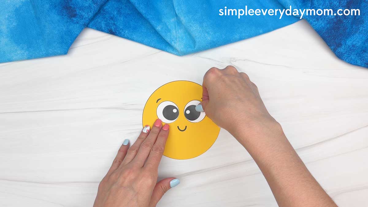 Hands gluing googly eyes onto a yellow sun cutout for a graduation craft activity