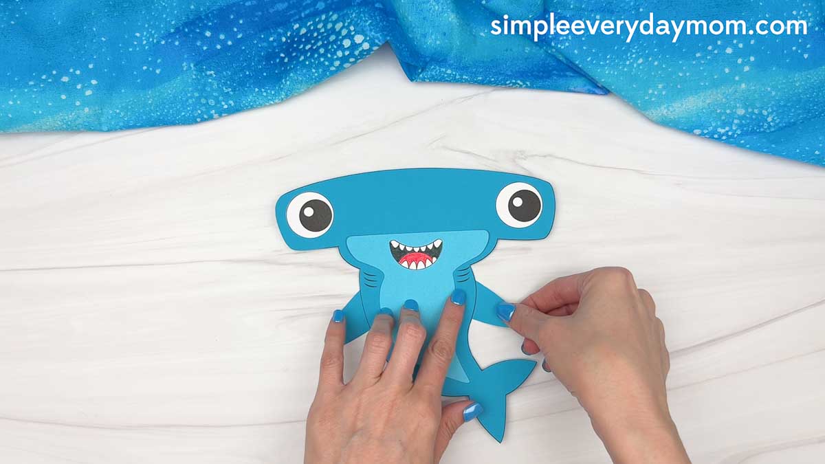 Hands attaching a side fin to a blue hammerhead shark craft during assembly.