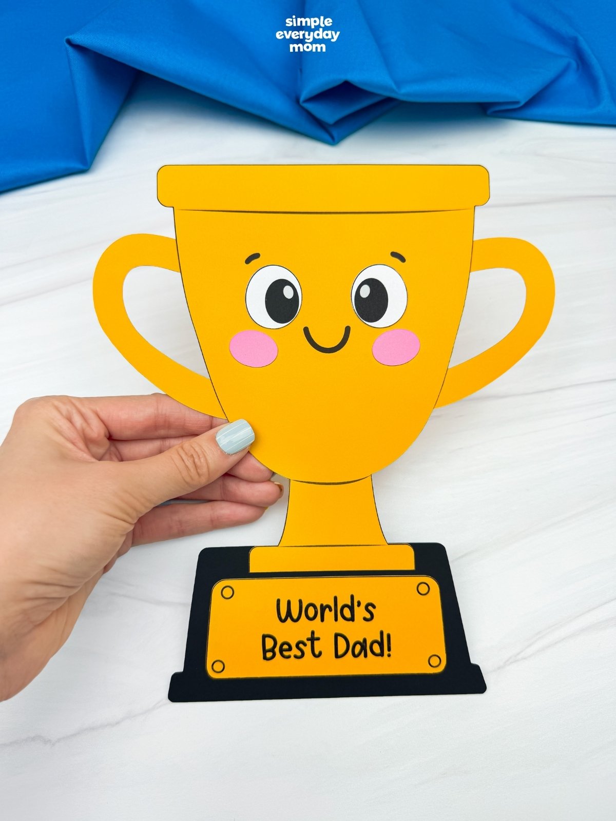 Hands holding a completed World’s Best Dad trophy craft with a smiling face and trophy handles.