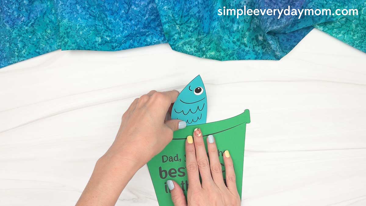Hands placing a paper fish into a green fishing bucket Father’s Day craft with a best catch message.