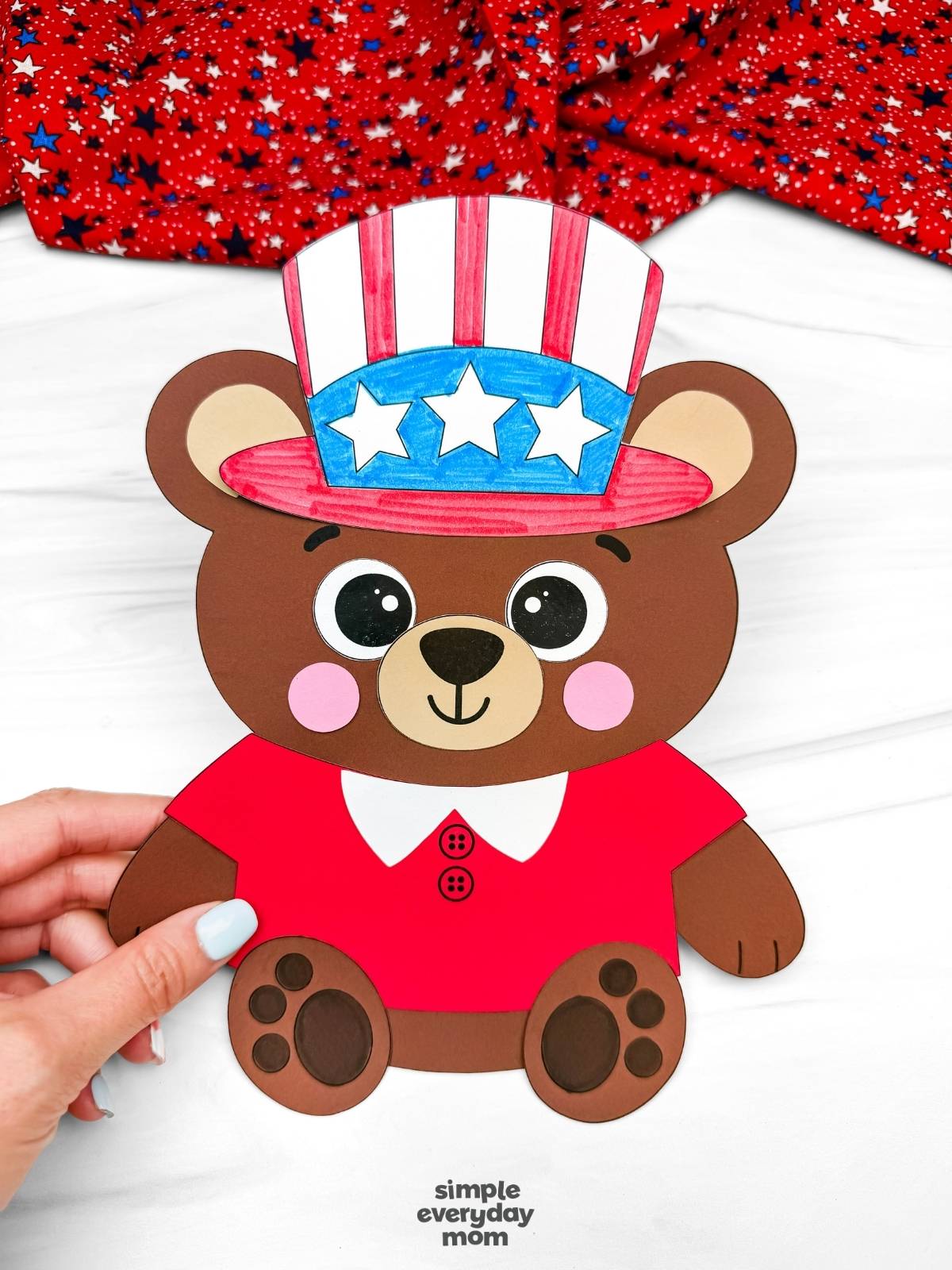 Hands holding a completed patriotic teddy bear paper craft with a red shirt and Uncle Sam hat for a Fourth of July kids activity