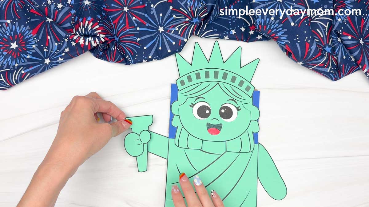 Hands attaching the Statue of Liberty torch arm piece to the side of the paper bag puppet craft.