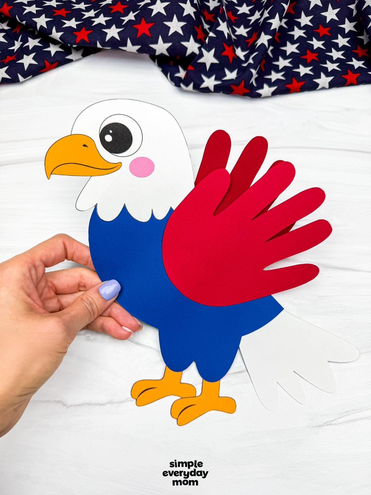 Hands holding a completed American eagle handprint craft with a blue body, red handprint wing, and white tail feathers.