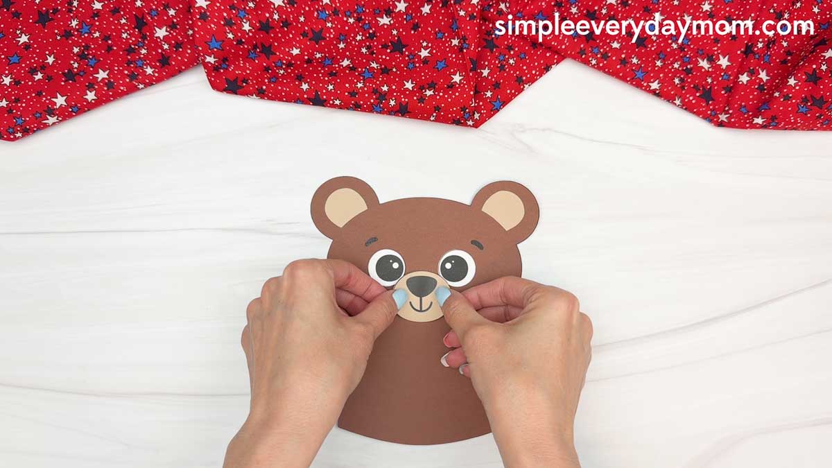 Hands placing the teddy bear muzzle onto a brown paper bear craft for a patriotic teddy bear activity