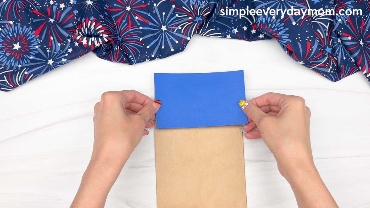 Hands placing a blue paper rectangle onto a brown paper bag to start the Statue of Liberty puppet craft.